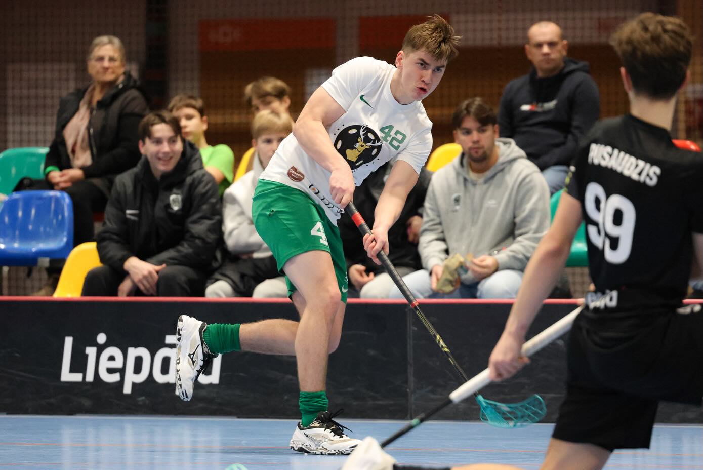 Florbola spēlētājs baltā kreklā un zaļos šortos sit bumbiņu ar nūju sporta zālē, skatītāji sēž tribīnēs.