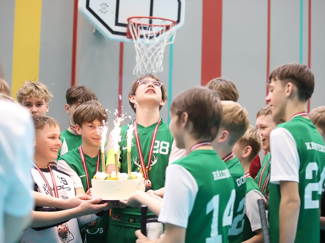 Bērnu florbola komanda svin jubileju sporta zālē ar kūku un svecēm pie basketbola groza.