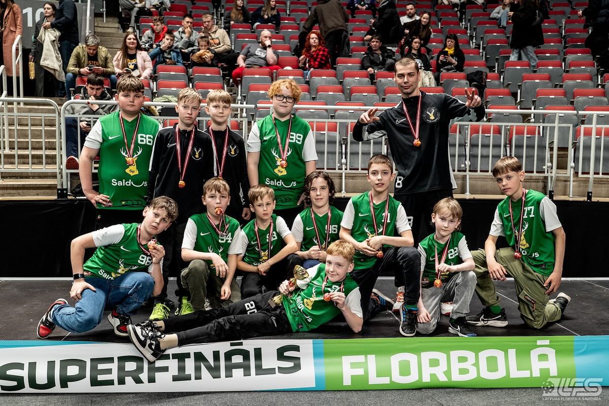 Bērnu florbola komanda ar medaļām un kausu pēc sacensībām stadionā ar skatītājiem tribīnēs.