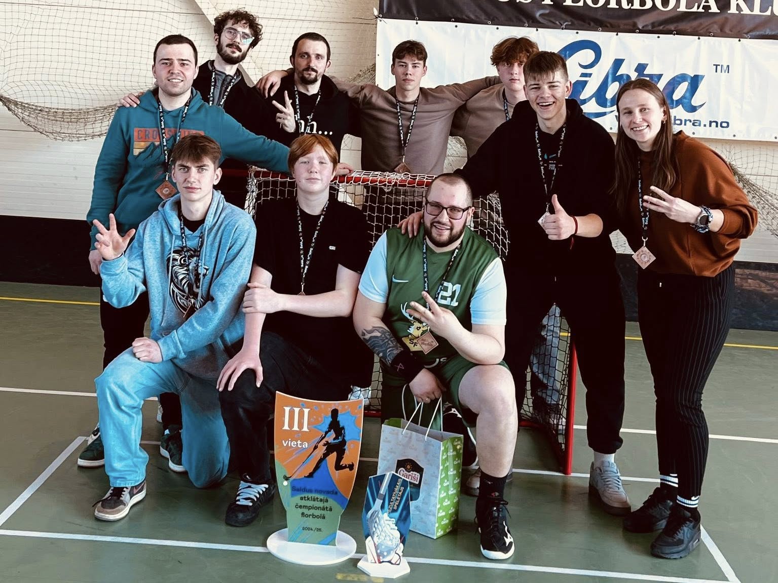 Deviņu jauniešu komanda sporta hallē ar medaļām un trofeju par 3. vietu florbolā.