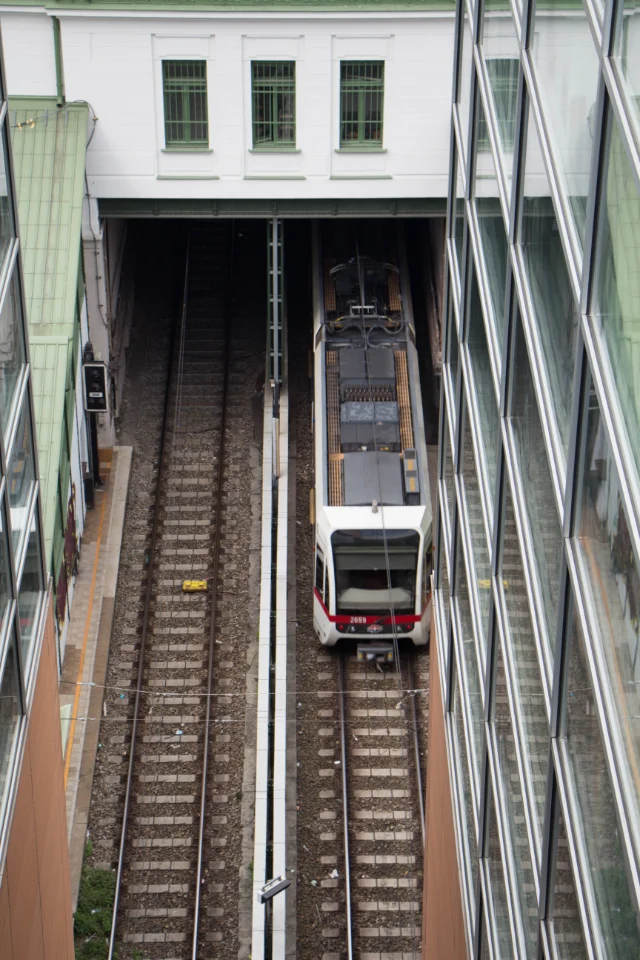 Straßenbahn fährt in einen Tunnel zwischen einem Glasgebäude und einem weißen Gebäude mit grünen Fenstern.