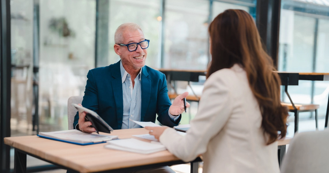 Smiling man in glasses and suit talks to woman across table in a modern office setting.