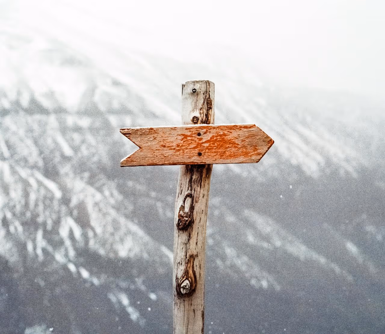 Weathered wooden arrow sign pointing to the right against a blurred mountainous background.