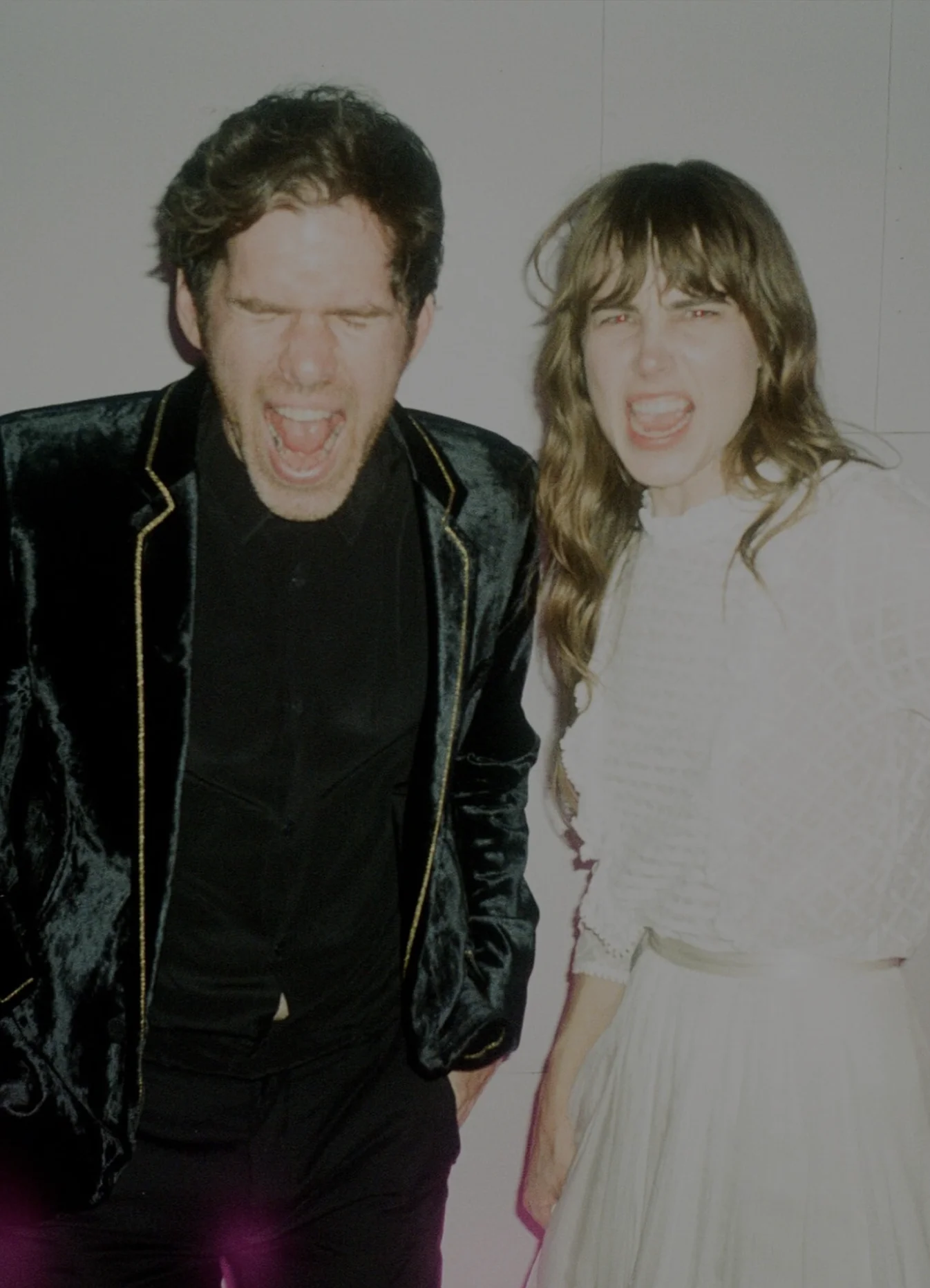 A candid photo of a man and woman screaming joyfully, standing side by side in stylish vintage outfits. The man wears a dark velvet jacket with gold trim, and the woman wears a white, long-sleeved dress with ruffled details. Photo by A Fist Full of Bolts.