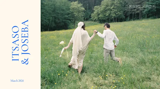 Couple holding hands and walking through a green meadow with yellow flowers surrounded by trees, the woman wearing a bridal veil and holding a bouquet, the man wearing a white shirt and light pants.