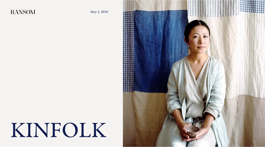 Portrait of a woman seated against a patchwork curtain backdrop in shades of blue and beige, holding a small ceramic bowl in her hands, wearing a light-colored layered outfit, with calm expression.