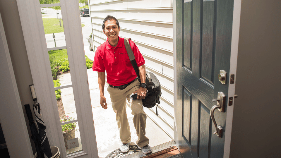 A Powerback Rehab to You therapist smiles as he walks through an open door of a patient's home. He is wearing a bright red polo shirt and khakis.