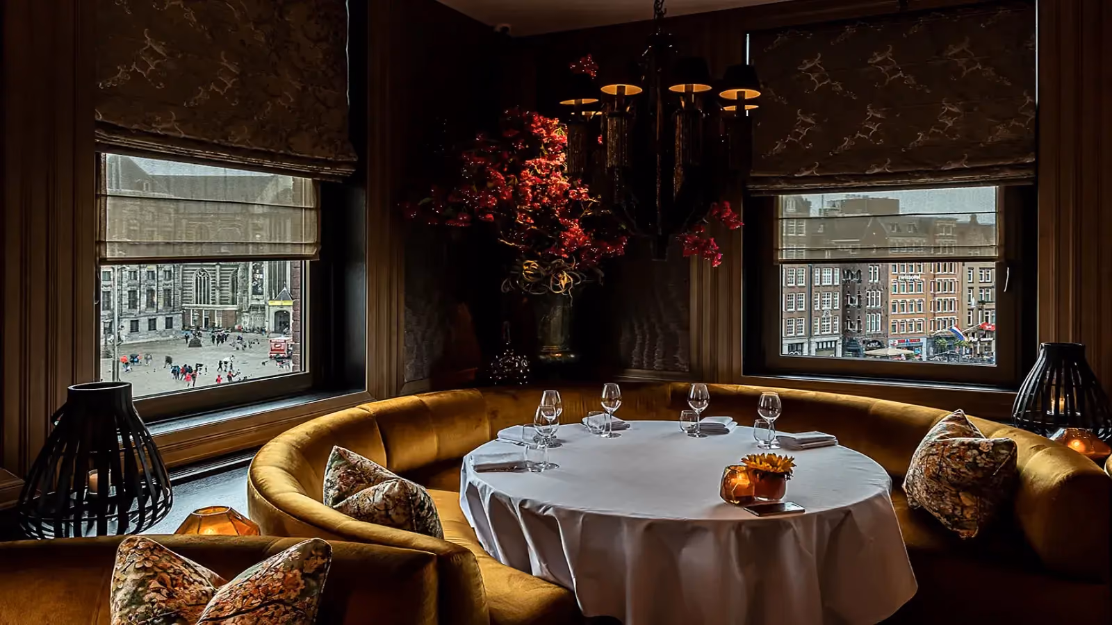 Cozy dining nook with mustard velvet seating, round white tablecloth table, and city views through windows.