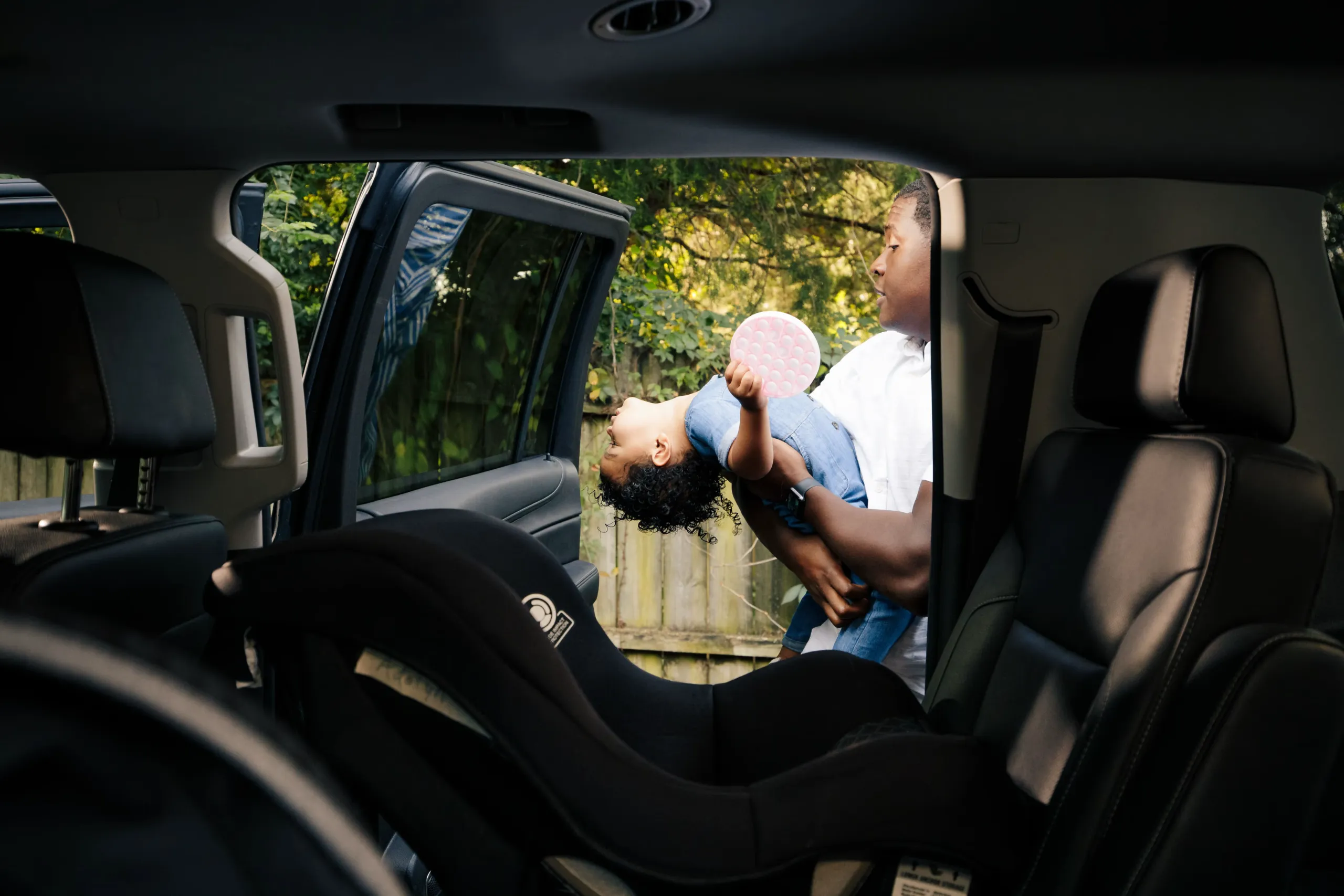 A small child trying to squirm out of his father's grasp, while attempting to put the child into their car seat