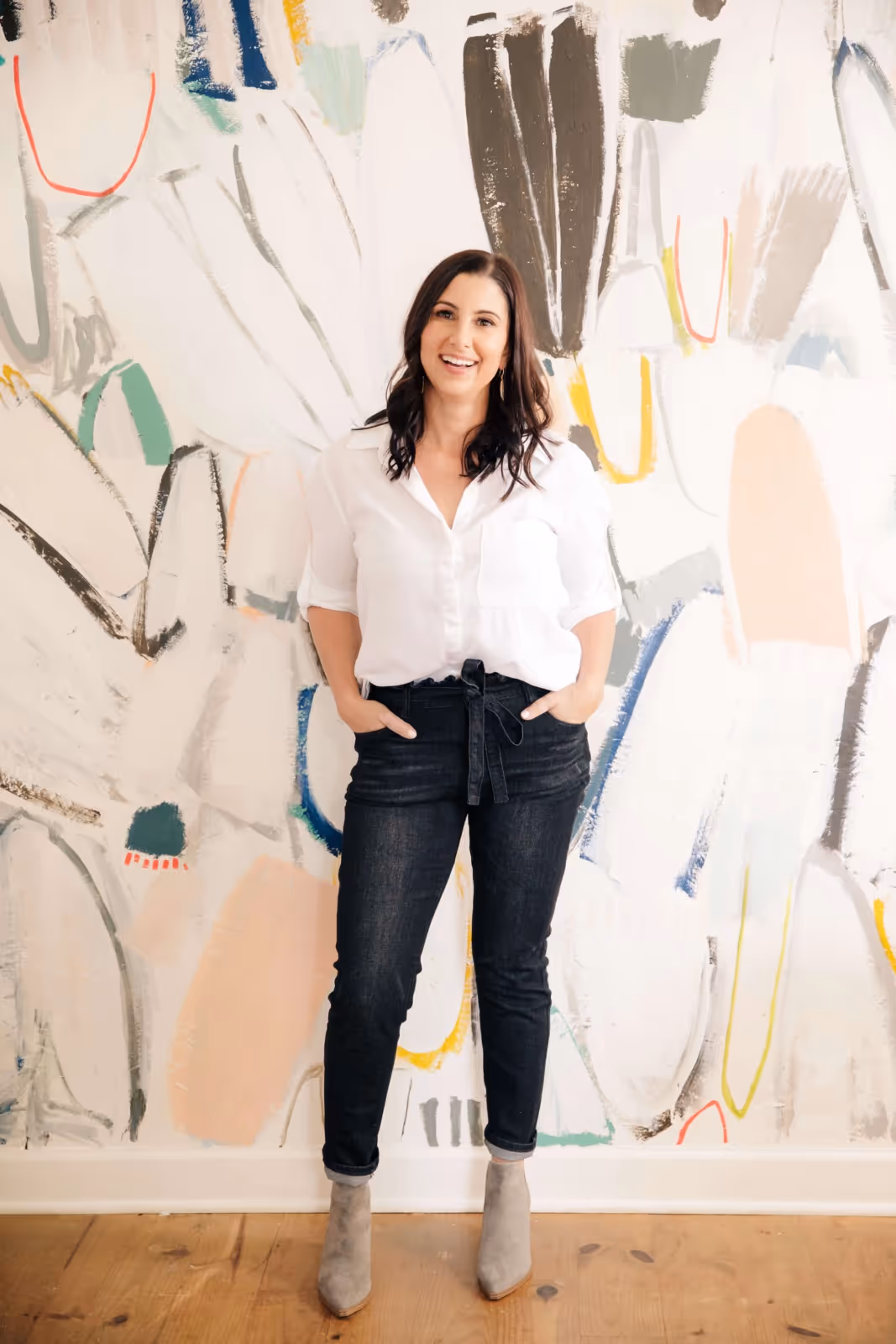 Smiling woman with dark hair wearing a white blouse, black jeans, and gray ankle boots standing against a colorful abstract wall.