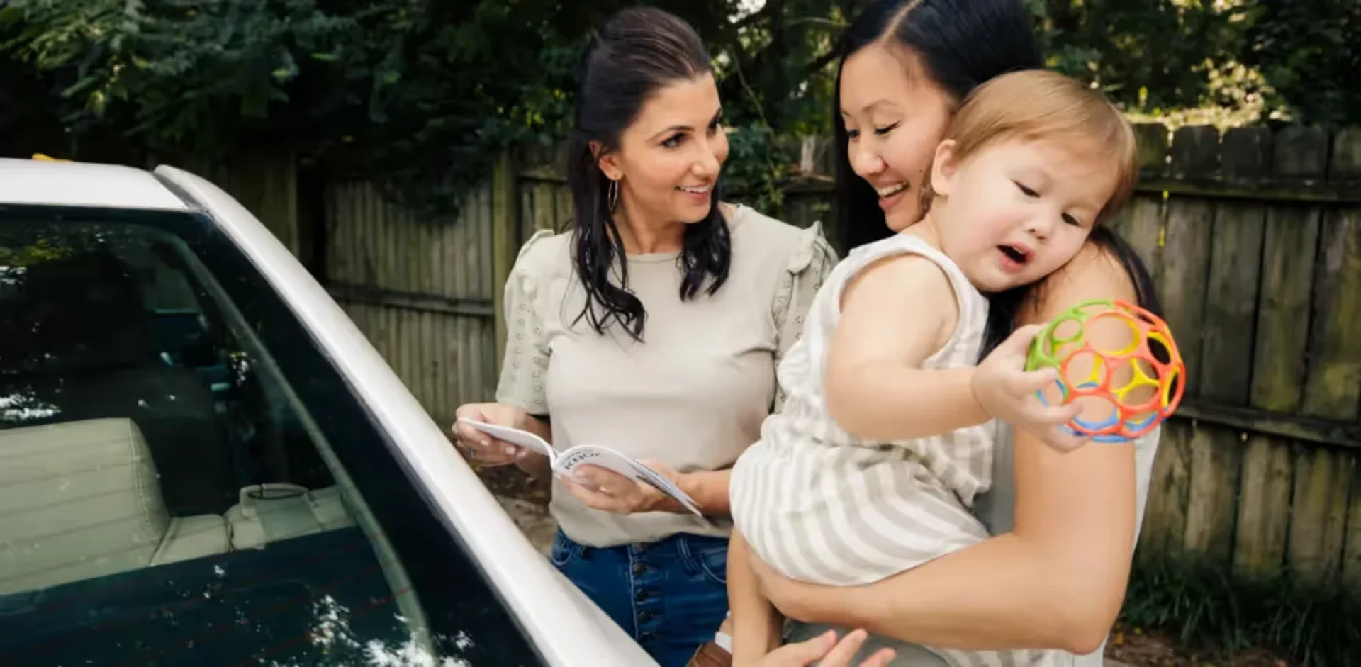 Woman holding toddler with toy talks to another woman reading car seat manual near a car.