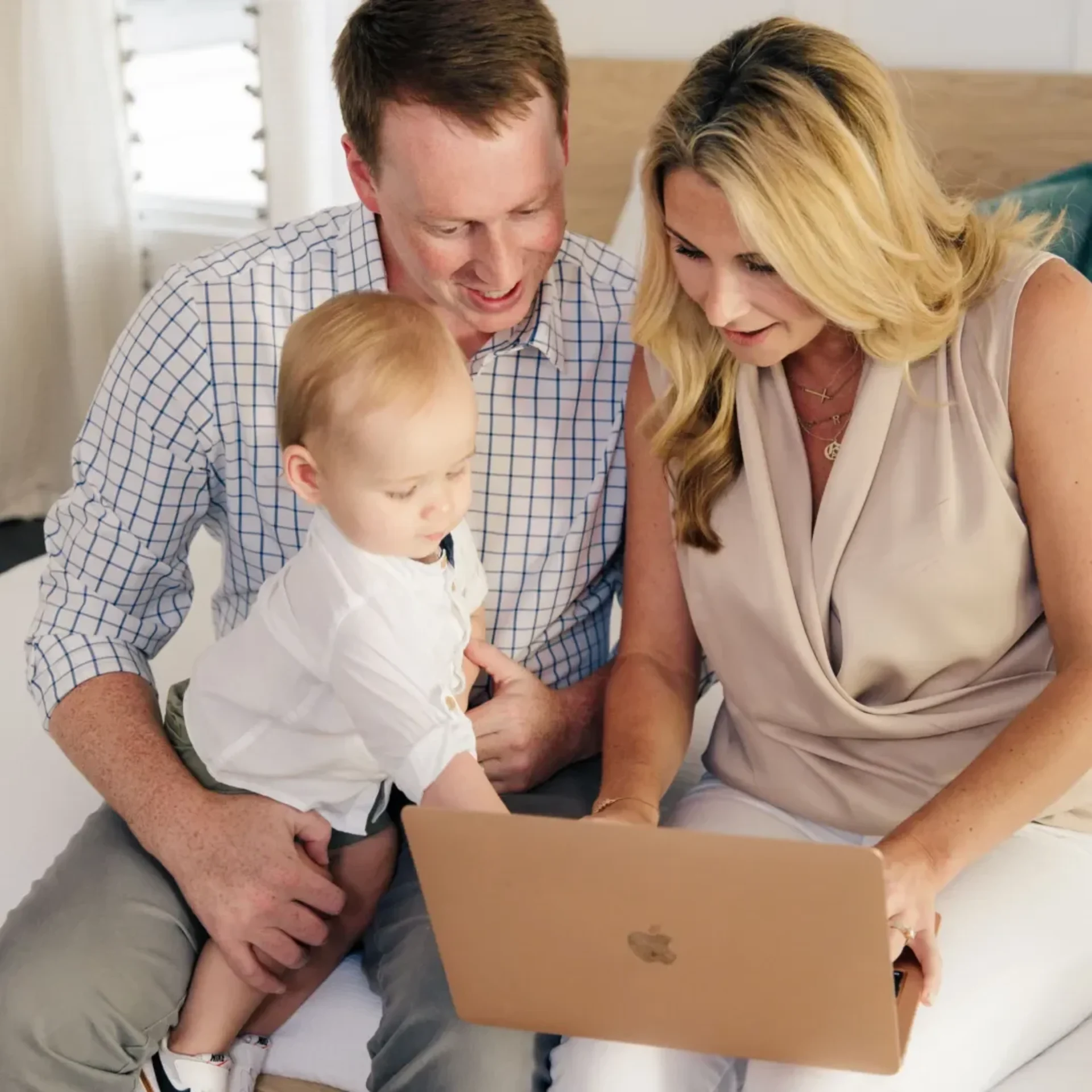 Parents and toddler sitting on a bed looking at a laptop together.