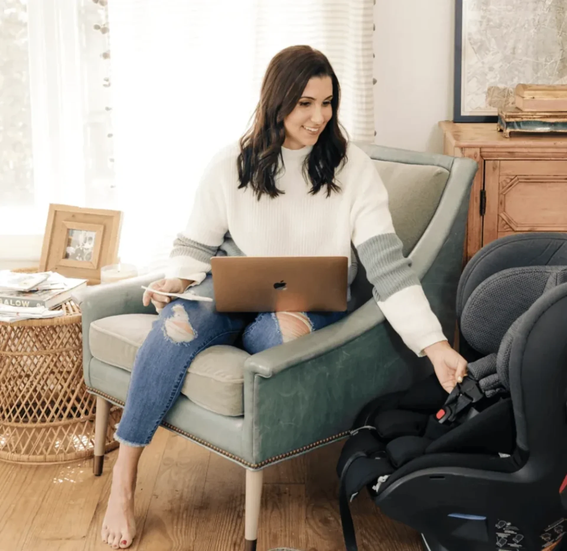 Woman sitting in a chair with a laptop on lap, reaching to adjust a black infant car seat nearby.