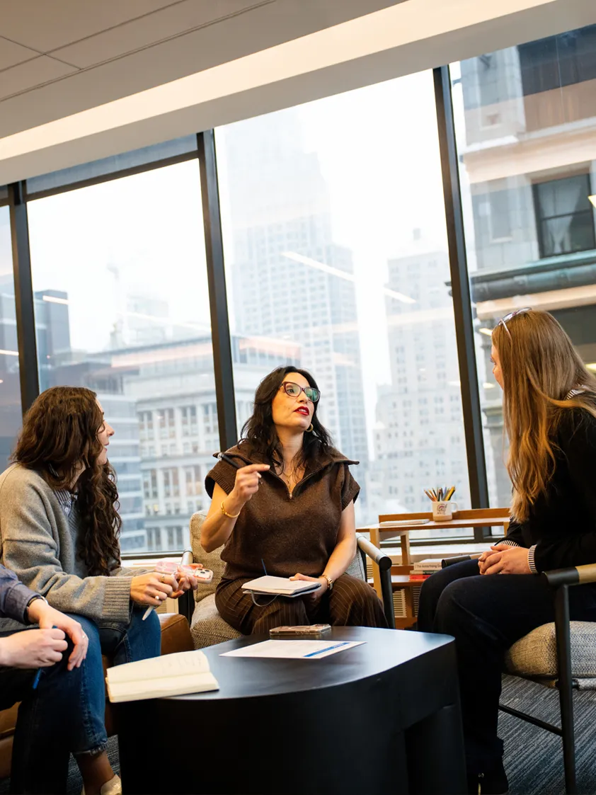 Three people seated in an office lounge having a team discussion with notebooks and papers on the table.