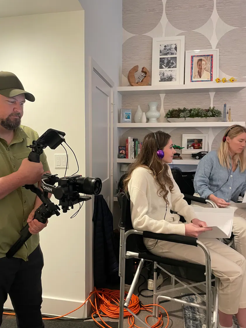 Camera operator filming two people during a behind-the-scenes video shoot, including one person seated in a wheelchair wearing headphones.