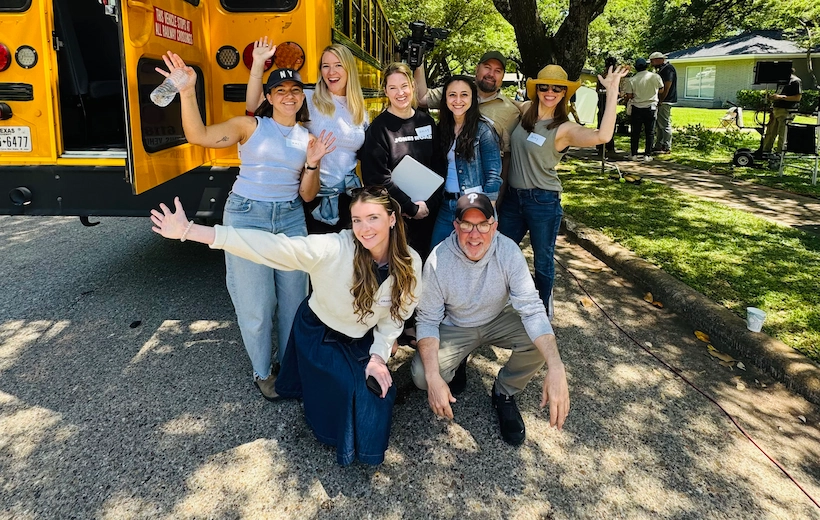 A group of eight colleagues pose enthusiastically in front of a yellow school bus outdoors on a sunny day. Six people stand in the back row waving and smiling, while two people crouch in front. They are casually dressed and appear to be on a team outing or work event.
