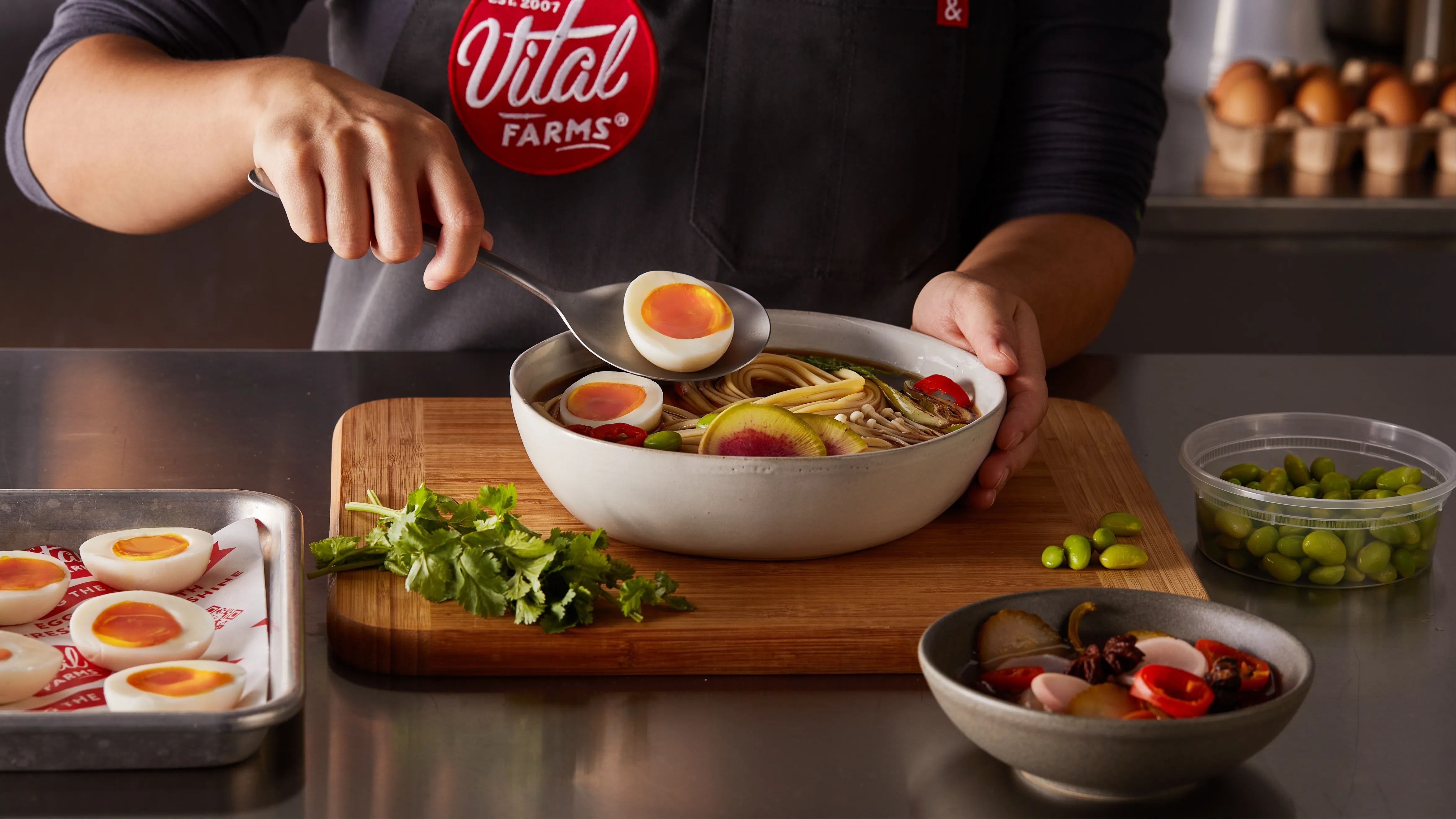 A person in a Vital Farms apron adds a halved soft-boiled egg to a bowl of ramen with noodles, vegetables, and herbs on a wooden cutting board, surrounded by ingredients and garnishes.
