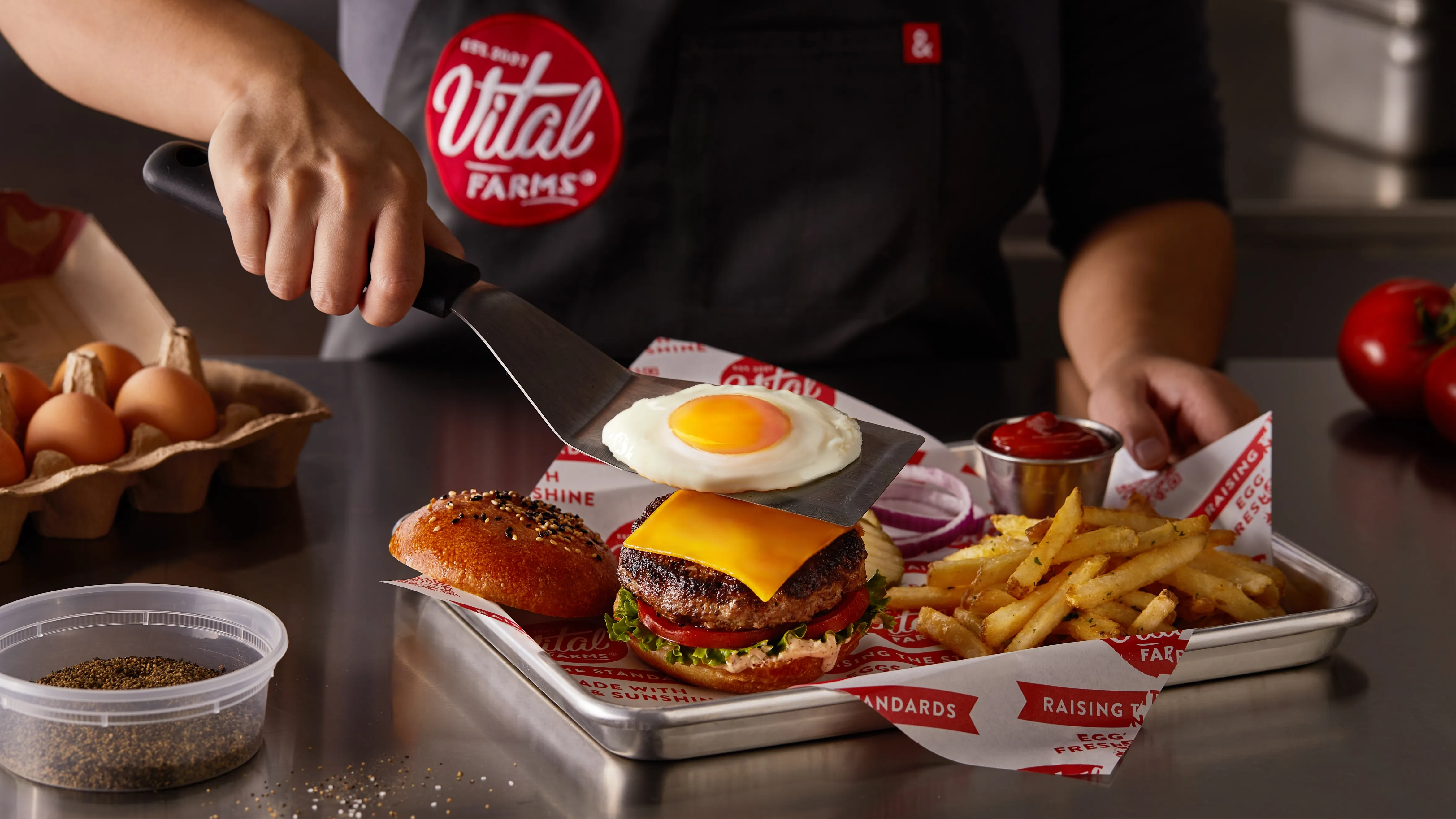 A person in a Vital Farms apron places a sunny-side-up egg onto a cheeseburger with lettuce on a tray, next to fries, ketchup, onions, and tomatoes, with eggs and pepper nearby.