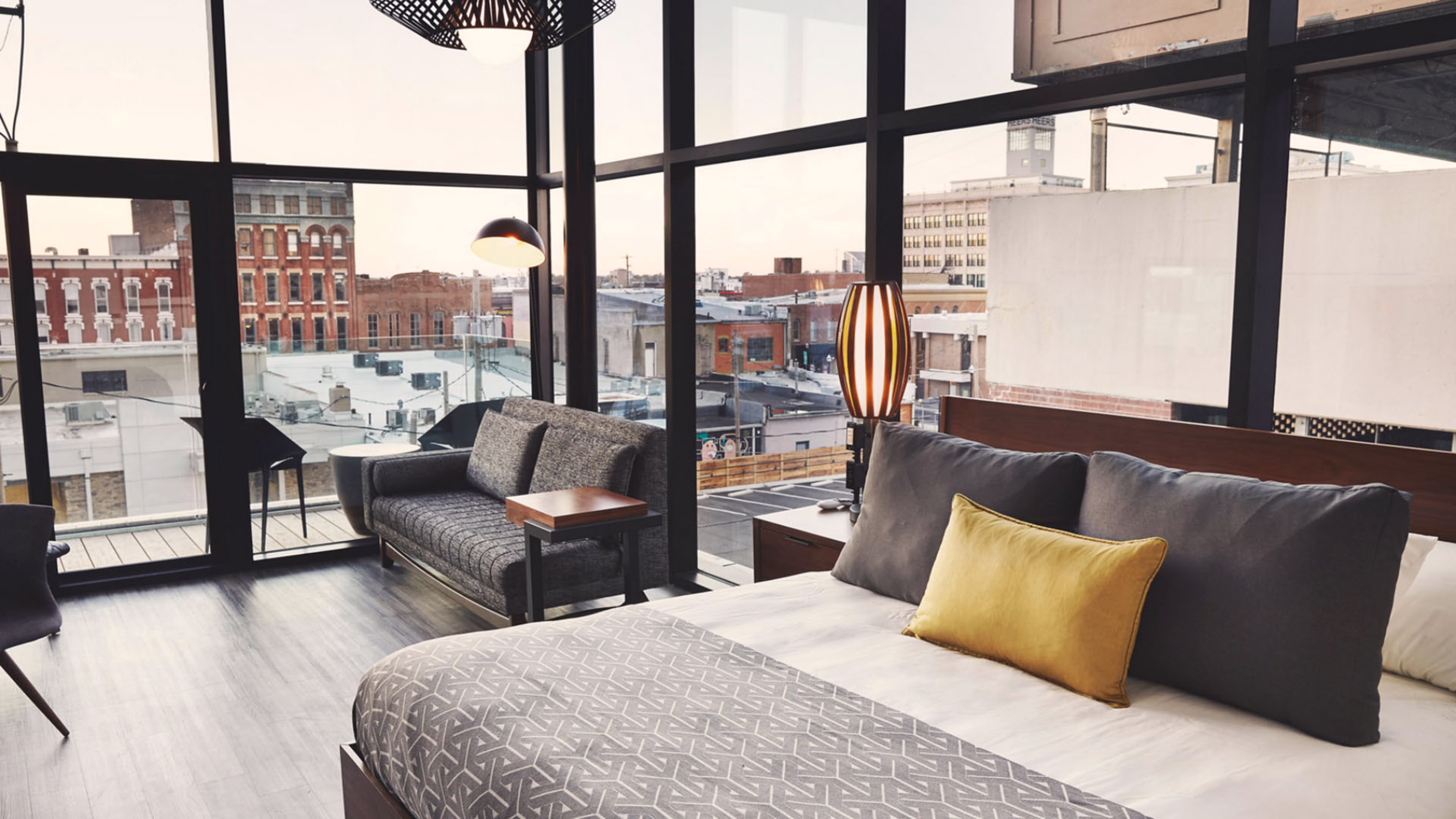 A modern hotel room with floor-to-ceiling windows overlooking a city skyline, featuring a bed with a yellow pillow and a grey sofa.