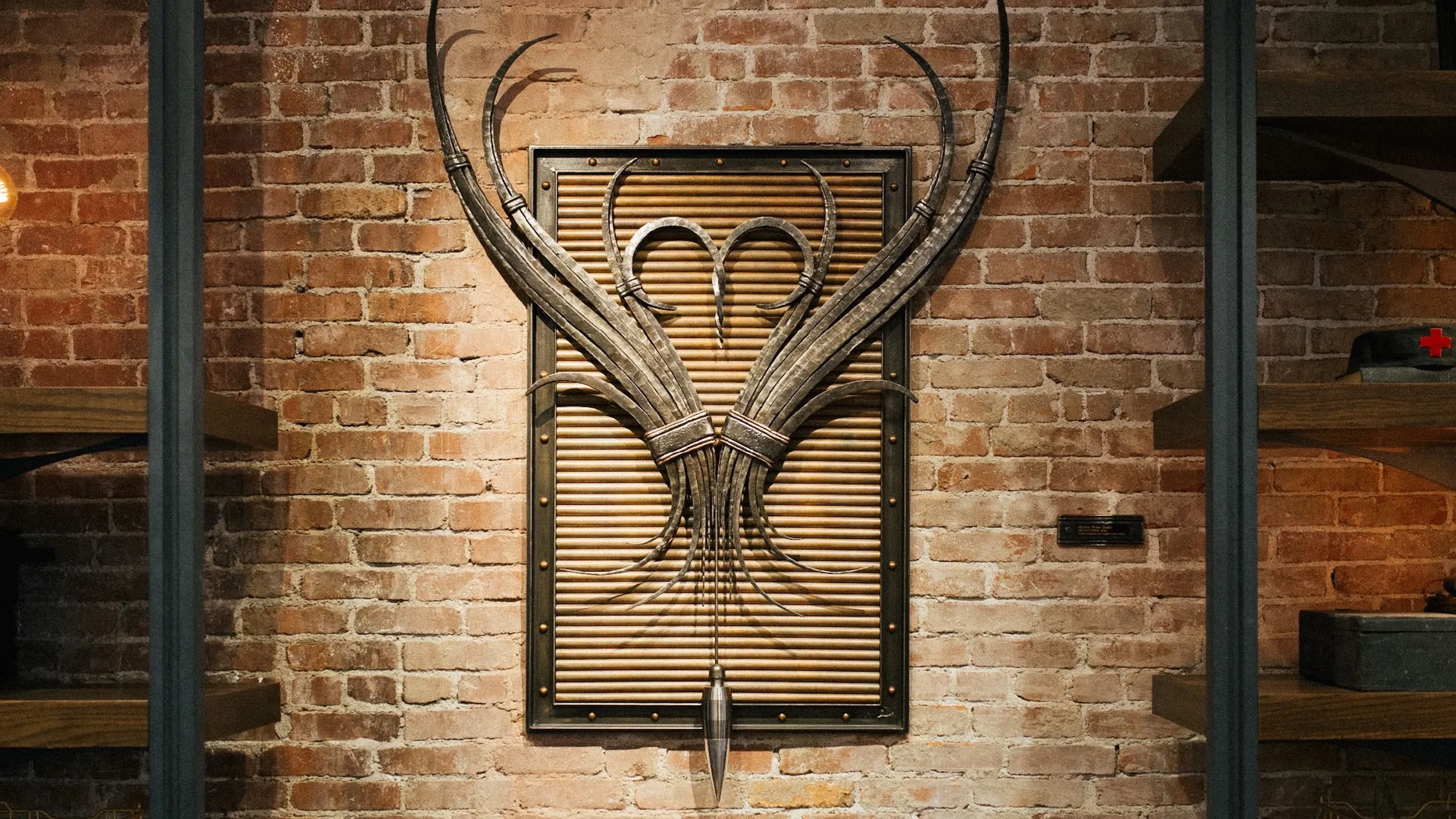 A decorative metal wall sculpture featuring symmetrical, curved blades and a central plumb bob, mounted on a brick wall.
