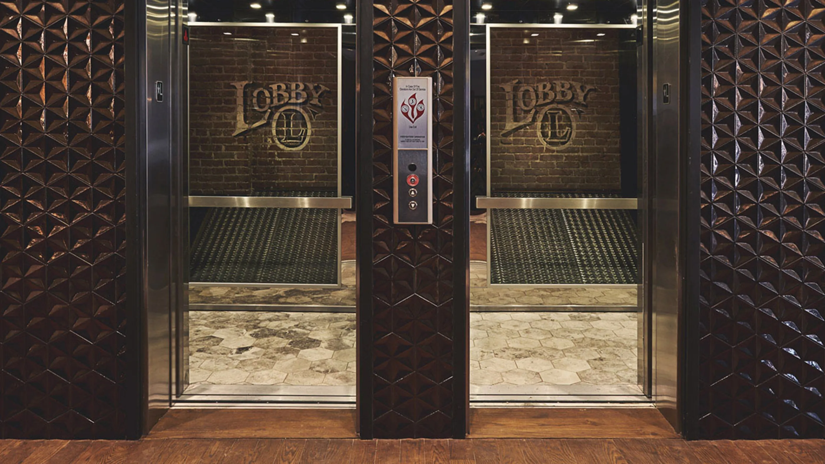 Two elevator doors side-by-side with a brick wall visible inside, featuring a LOBBY logo and an emergency call panel between them.