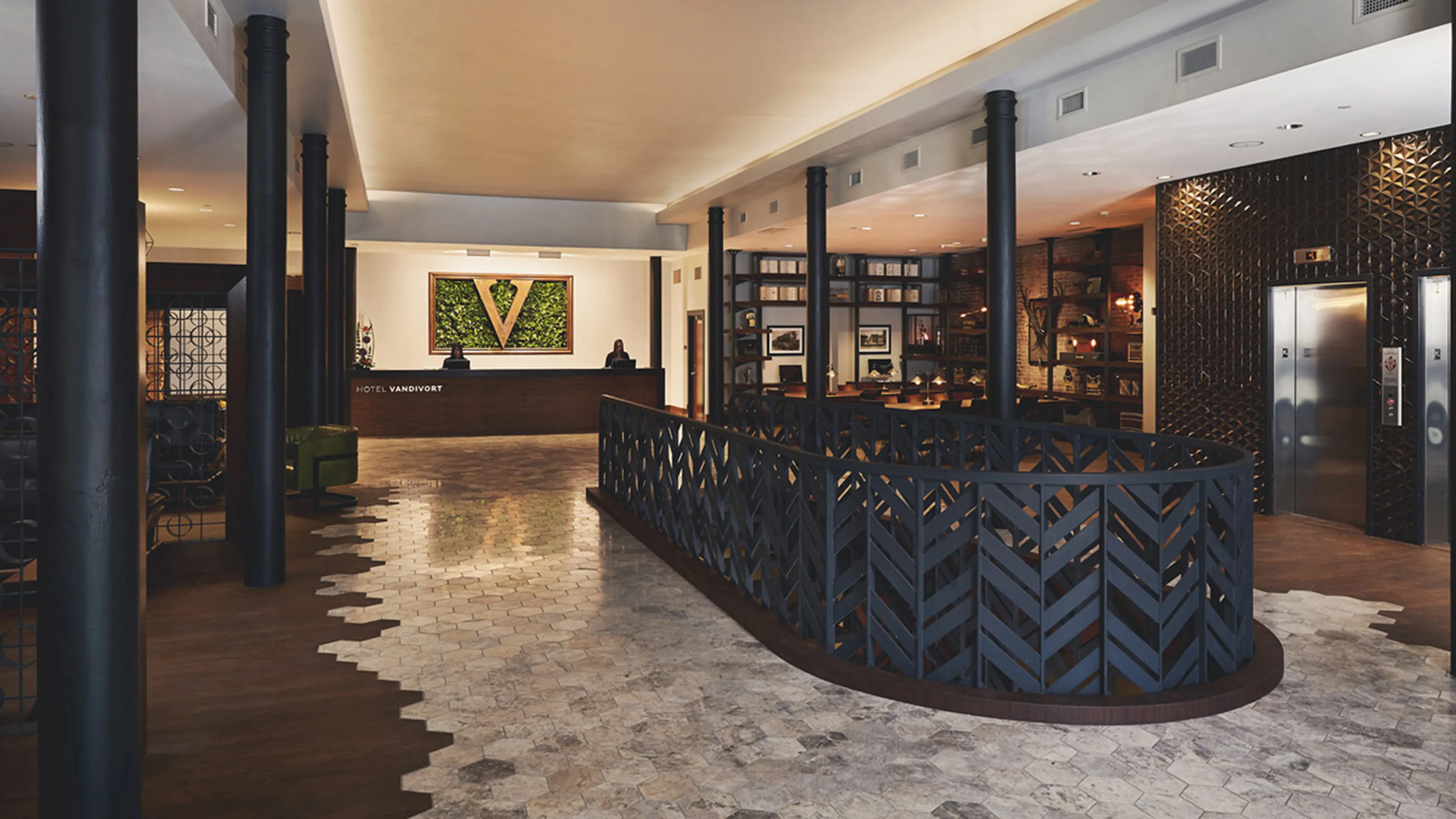 Hotel Vandivort lobby featuring a dark wood reception desk with a large V logo, a black geometric metal railing, and a light-colored hexagonal tile floor.