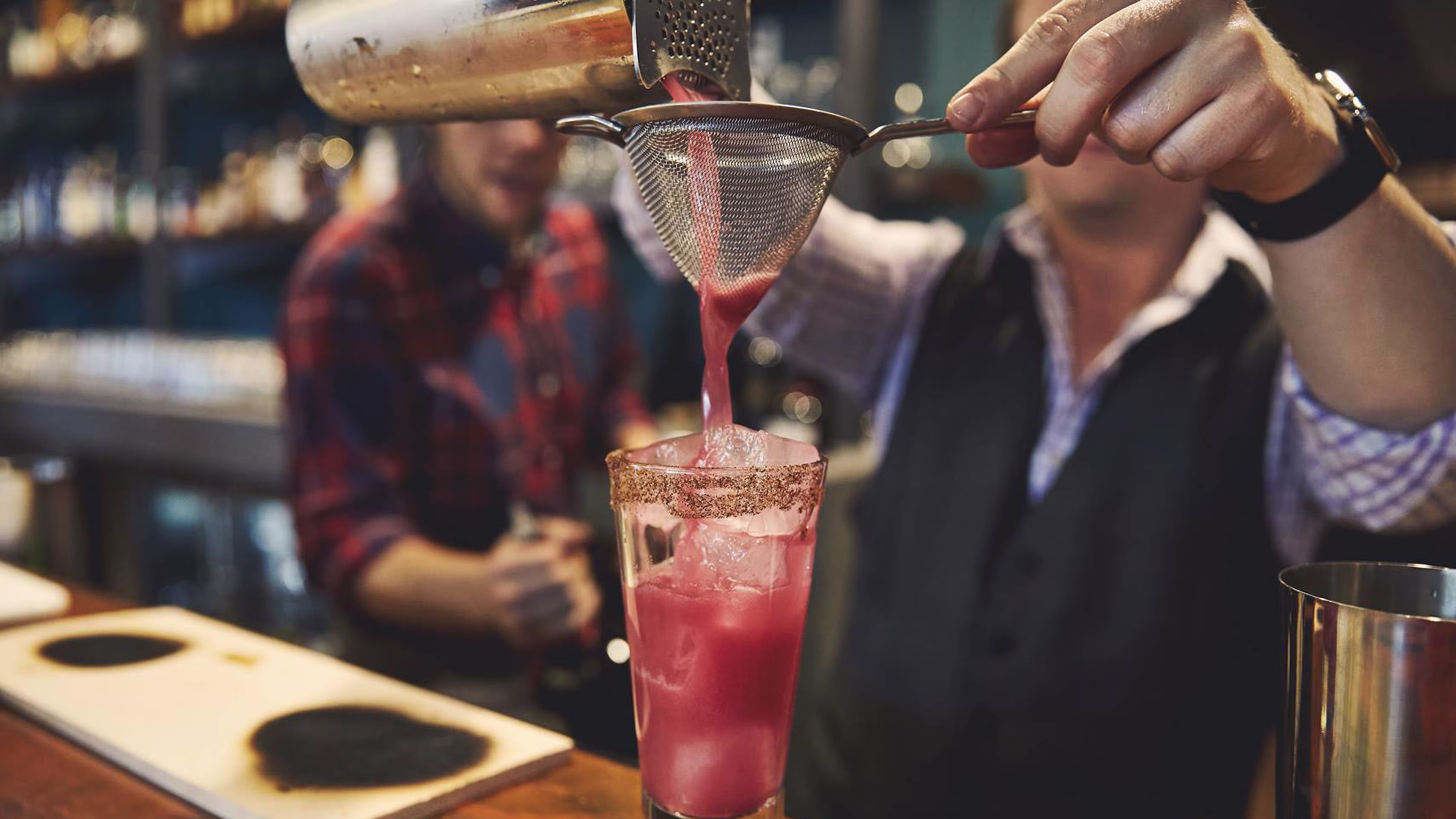 A bartender strains a pink cocktail into a rimmed glass filled with ice.