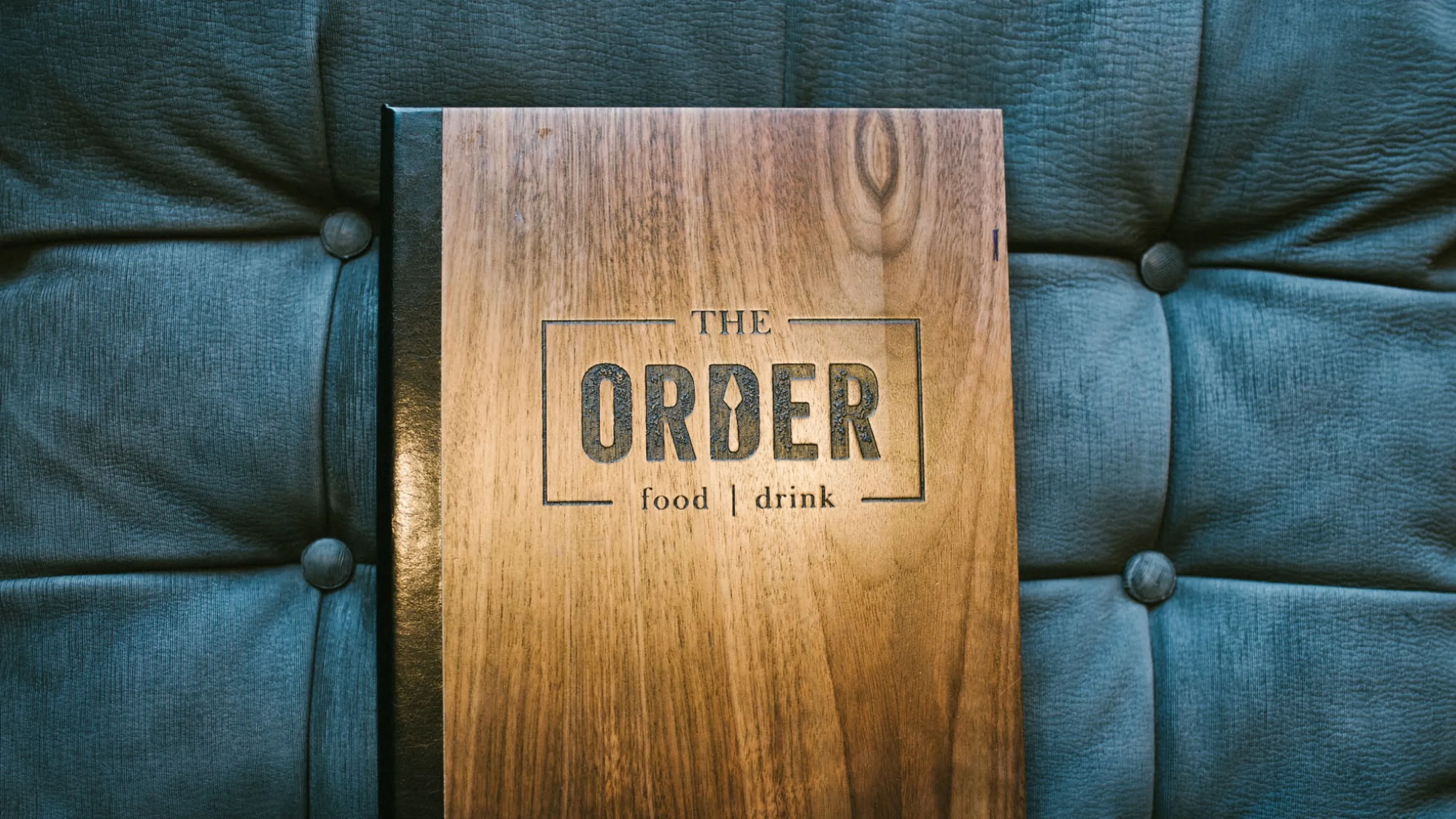 A wooden menu cover labeled The Order food | drink resting on a tufted, dark blue surface.