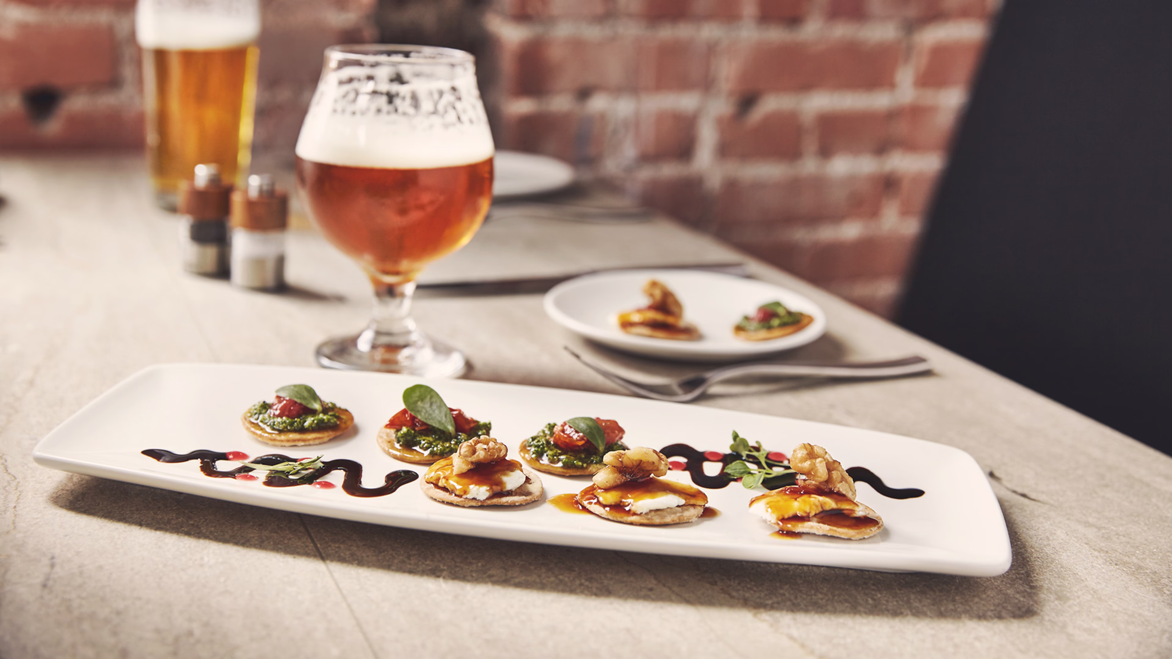 A rectangular serving plate featuring small appetizer crackers topped with pesto, cheese, fruit, and walnuts, accompanied by a glass of beer.
