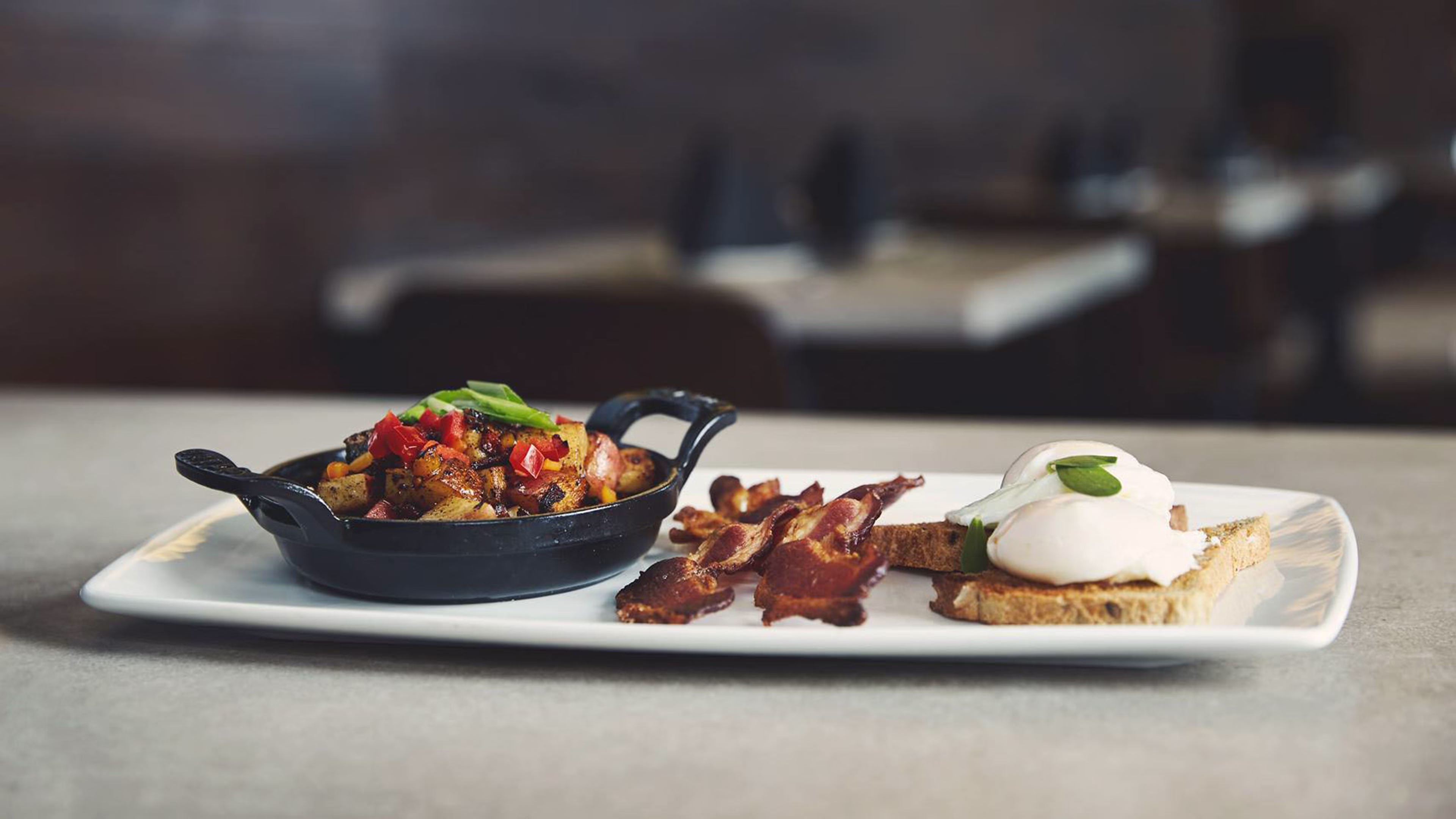 A breakfast plate featuring a small skillet of roasted potatoes, crispy bacon, and two poached eggs on toast.