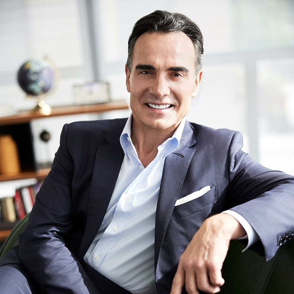 Smiling middle-aged man in a navy suit and light blue shirt seated in a modern office setting.