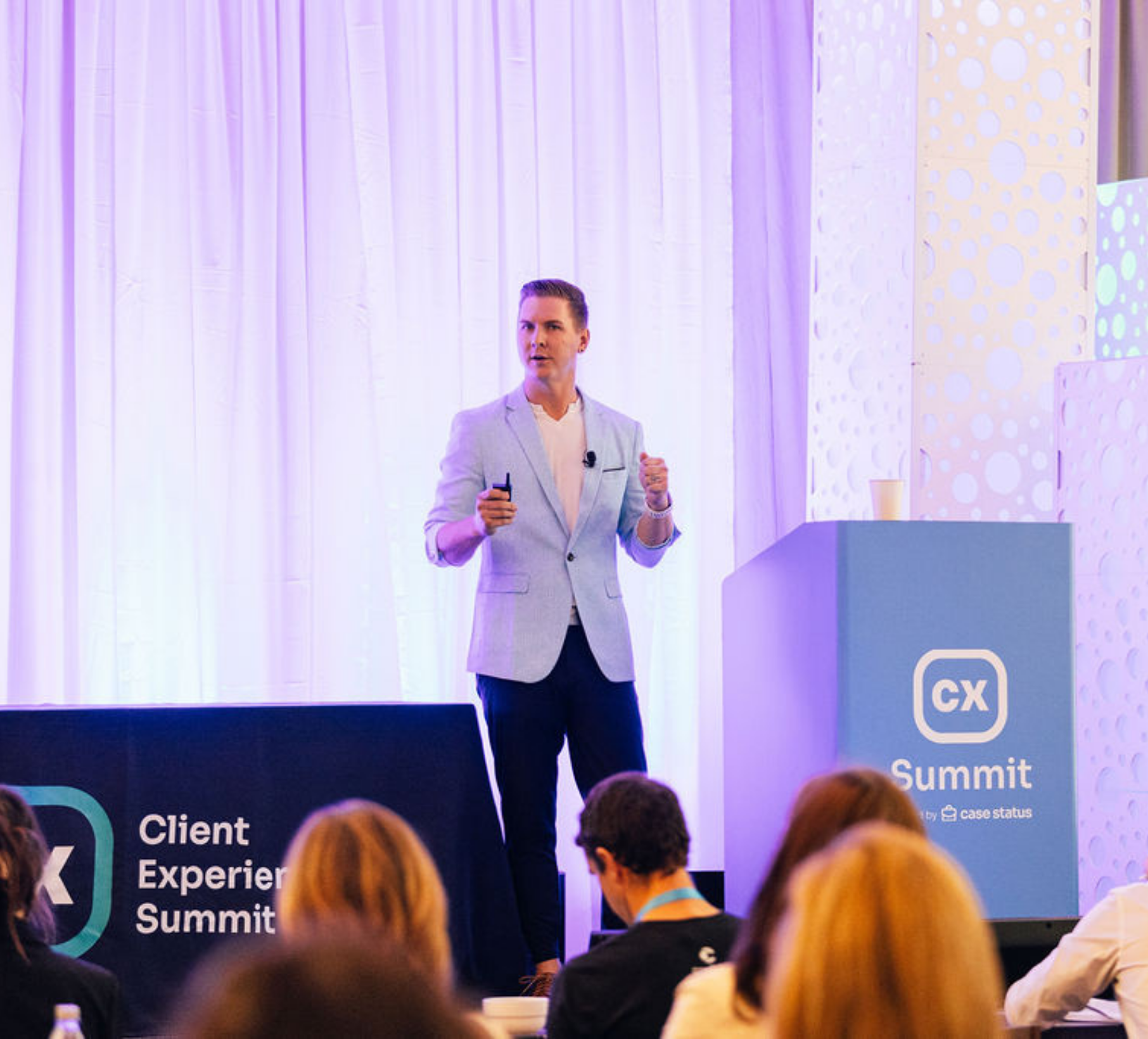 Presenter in light blue blazer speaking at Client Experience Summit on stage with audience in foreground.