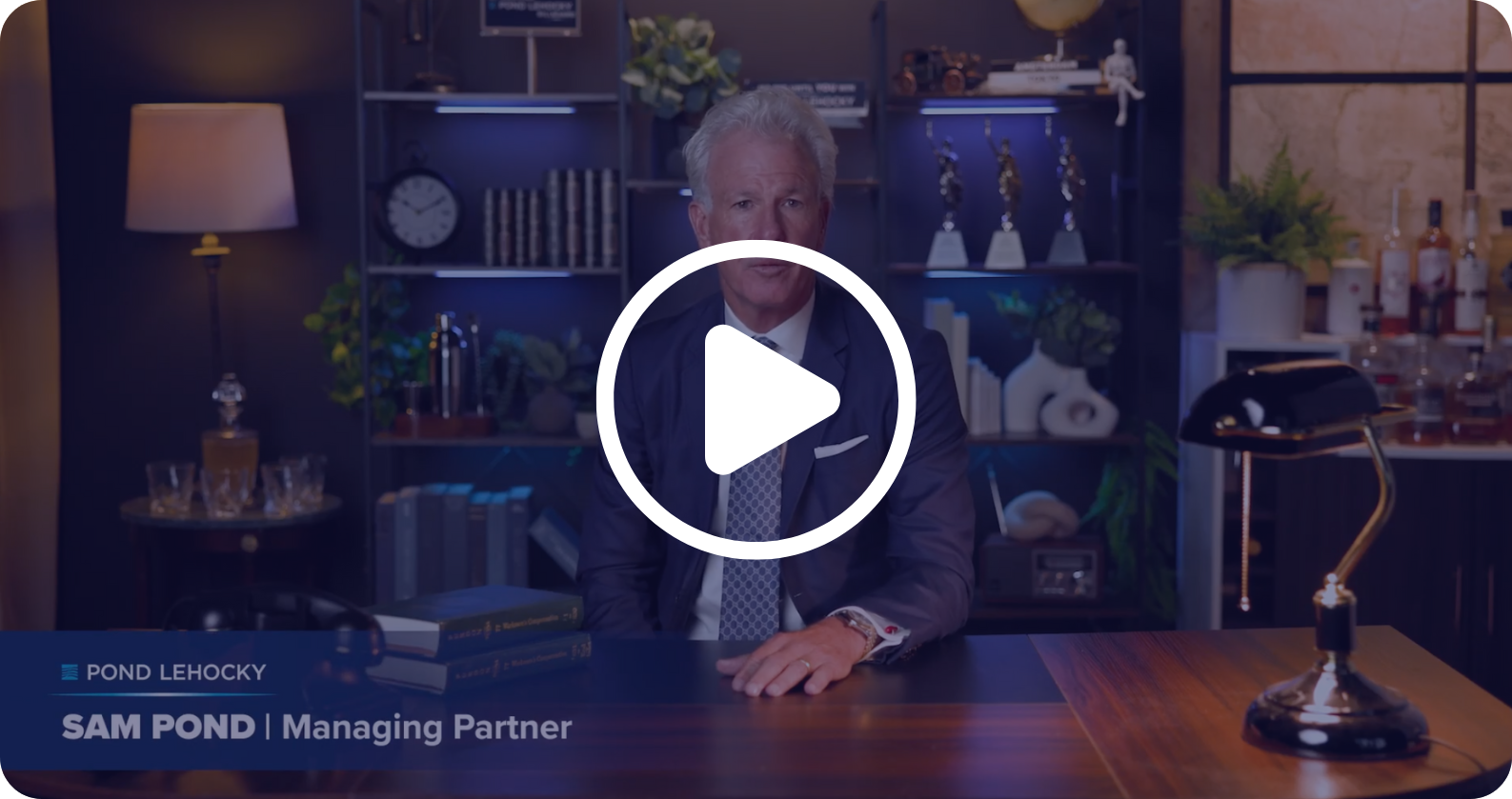 Man in suit sitting at wooden desk in an office with shelves, trophies, lamp, and bar setup in the background, identified as Sam Pond, Managing Partner at Pond Lehocky.