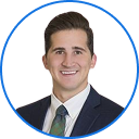 Smiling young man in a suit and tie with neatly styled brown hair against a white background.