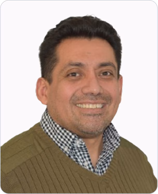 Smiling man with dark hair wearing a green sweater over a checkered shirt against a plain white background.