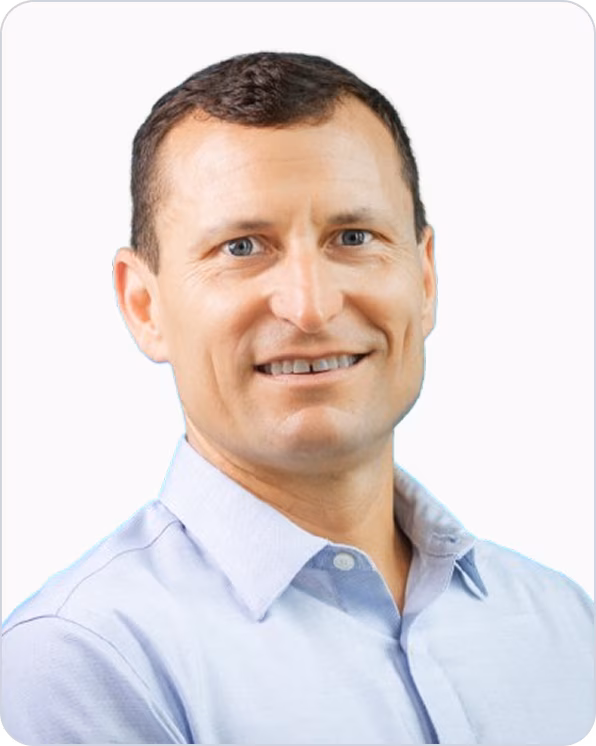 Smiling man with short dark hair wearing a light blue button-up shirt against a white background.