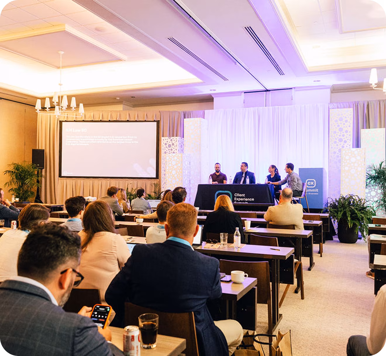 Audience attending a panel discussion at a conference with speakers seated at a table on stage and a presentation screen in the background.