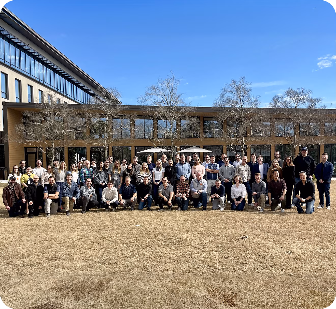 Large group of men and women wearing matching blue, teal, and light green t-shirts posing for a team photo against a plain light gray background.