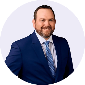 Smiling man with beard wearing a blue suit, white shirt, and striped blue tie on a light background.