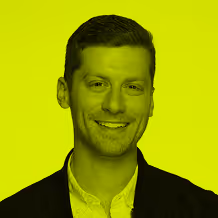 Smiling man with short hair wearing a white shirt and dark jacket against a yellow-green background.