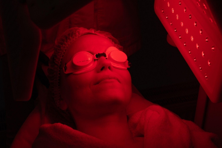 Person receiving red light therapy facial treatment under LED panel in dark room