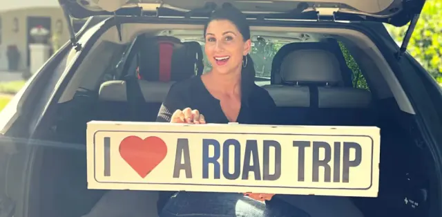 A woman sitting in the back of a car holding a road trip sign