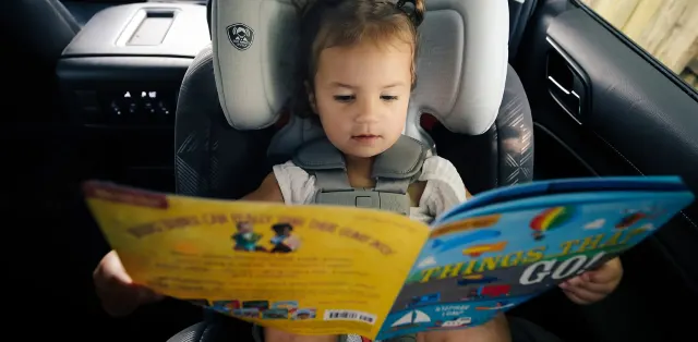 A little girl sitting in a car seat reading a book