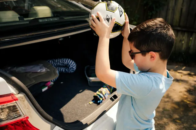 A young boy holding a soccer ball in the trunk of a car