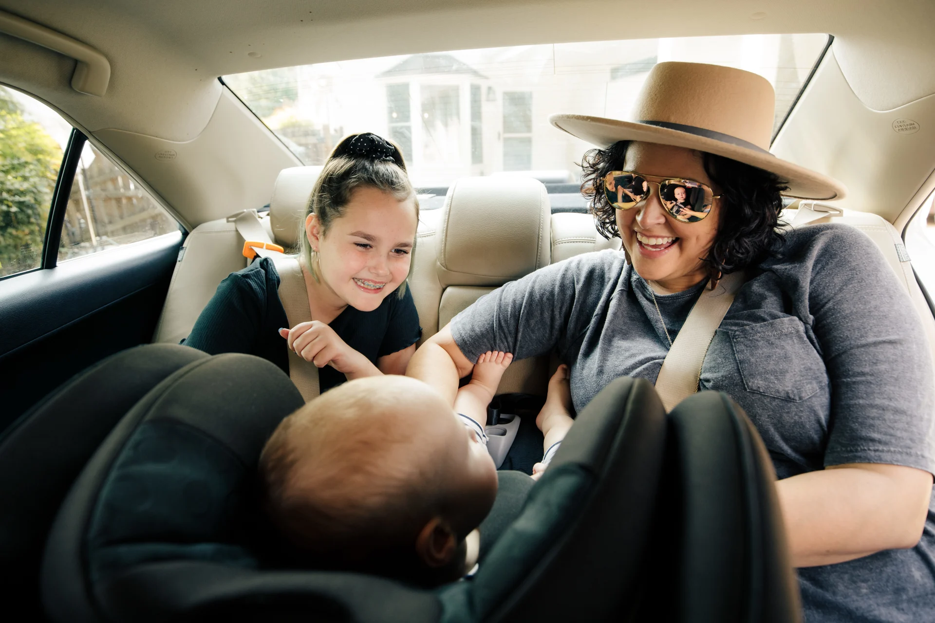 Family taking selfie in car backseat during road trip, woman wearing sunglasses and hat smiling with two children