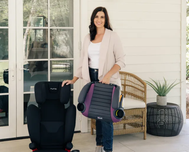 Smiling dark-haired woman on a white farmhouse porch holding a purple Chicco booster seat beside a black Nuna car seat