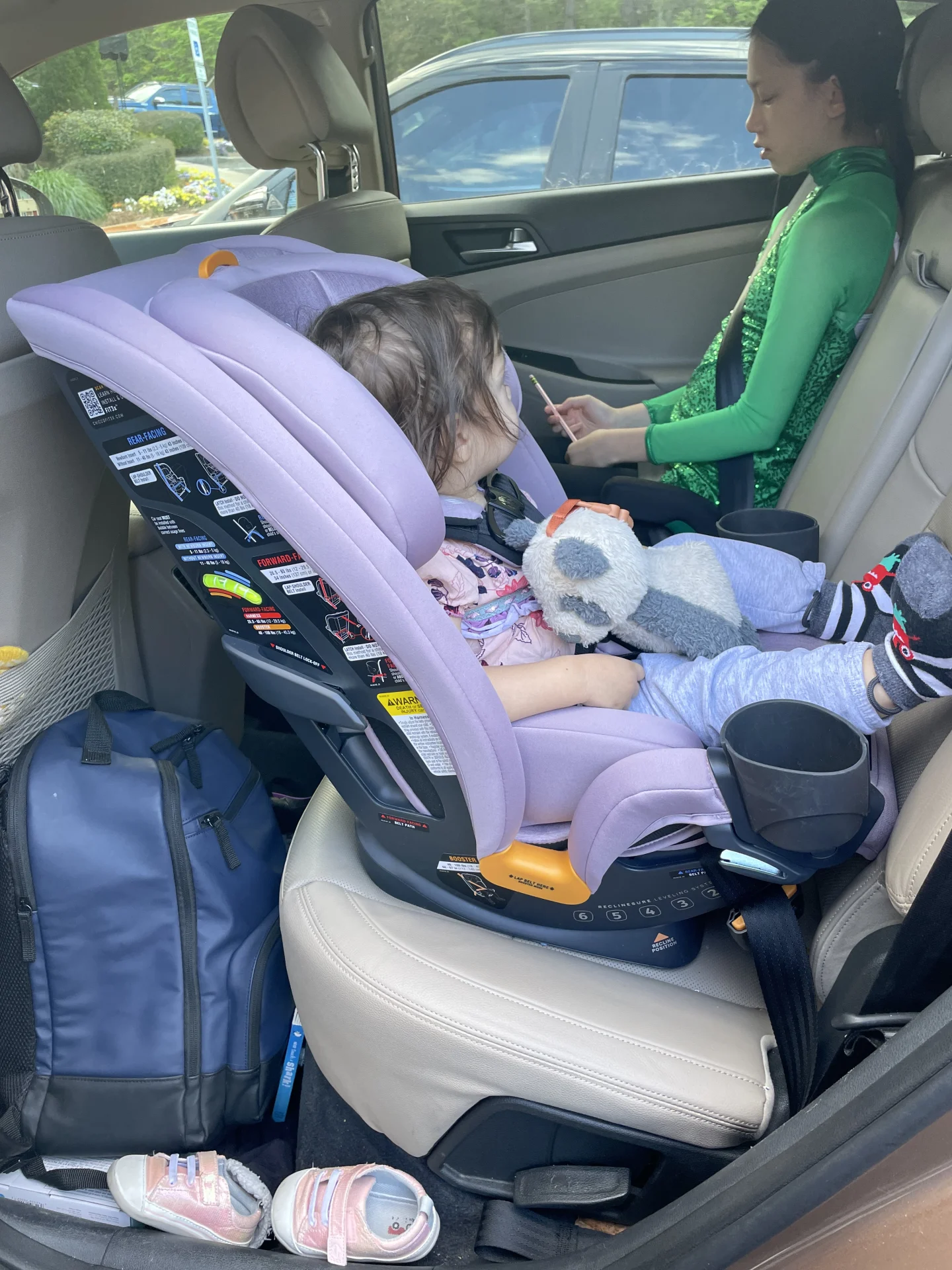 Toddler sitting in a lavender Chicco Fit3x convertible car seat in rear-facing position holding a stuffed animal in a vehicle