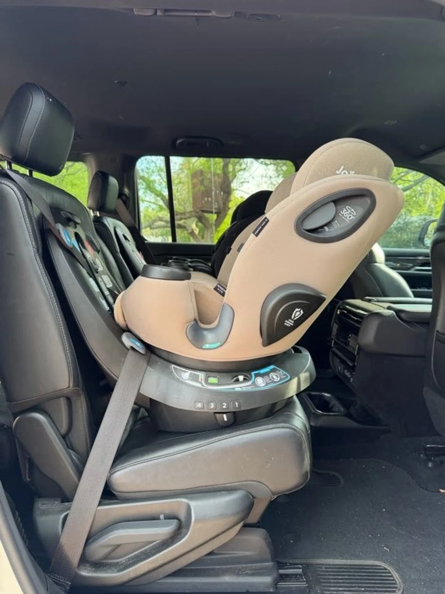 Joie Spin SI convertible child car seat in matcha tan installed and rotated in black leather SUV rear seat with trees visible