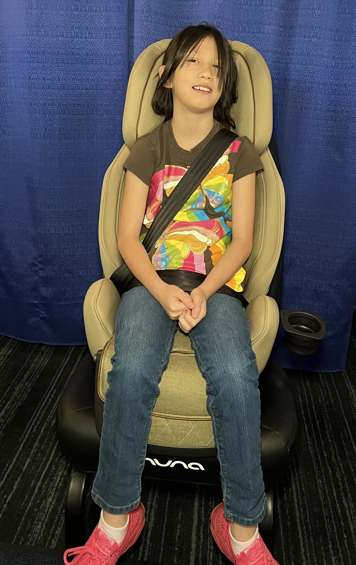 Smiling child with dark hair wearing a colorful graphic tee and jeans seated in a tan Nuna Exec booster car seat with seatbel
