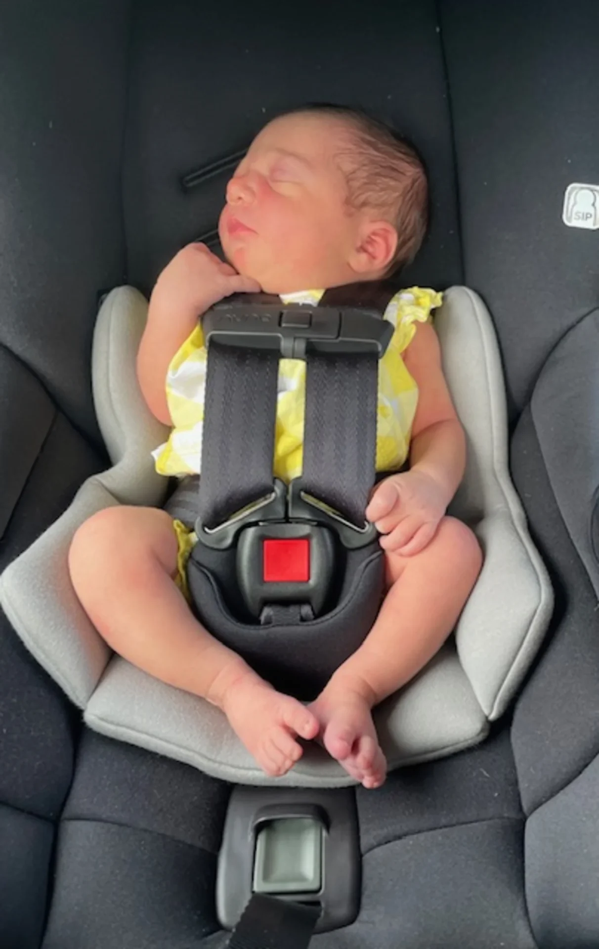 Peaceful sleeping newborn baby in yellow outfit securely buckled into a Nuna Pipa RX infant car seat with grey padding